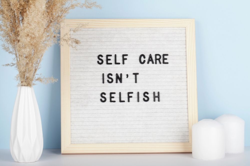 A wooden framed  board on a blue background displaying the inspirational message Self care isn’t selfish-
